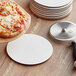 A 6" white pizza on a wood table next to a white corrugated pizza circle and a pizza cutter.