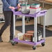 A woman pushing a purple Choice utility cart with containers on it.