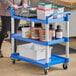 A woman using a blue Choice utility cart with three shelves to hold blue and white food containers.