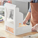 A person wearing gloves and a brown apron cutting an orange frosted cake in a white Baker's Mark bakery box.