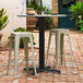 A black Lancaster Table & Seating Excalibur outdoor table base with white stools on a brick patio.