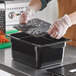 A person in gloves using a black plastic Vigor food tray with a lid and drain tray.
