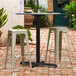 A black Lancaster Table & Seating Excalibur outdoor table base with bar stools on a brick patio.