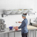 A woman in a blue uniform mixing food on a white Baker's Mark wall-mounted ingredient bin shelf.
