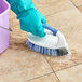 A gloved hand using a Quickie iron scrub brush to clean a tile floor.