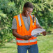 A man in an Ergodyne orange surveyor vest holding a clipboard and looking at a piece of paper.