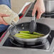 A hand cooks asparagus in a Vollrath stainless steel pan on a stove.