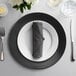 A black 10 Strawberry Street lacquer charger plate with a white napkin folded on it and silverware on a table.