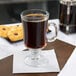 A Libbey Irish glass coffee mug filled with brown liquid on a table with a napkin and cookies.