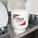 A white container with a red label of Noble Chemical Power Block Concentrated Solid Dish Machine Detergent on a metal shelf above plates.