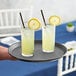 A gray non-skid round serving tray holding two glasses of lemonade with a straw on a table.