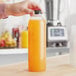 A hand holding a 32 oz. round clear PET juice bottle with orange liquid inside.