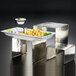 A tray of food on an American Metalcraft stainless steel riser with a white container on top.