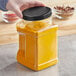 A hand holding a 64 oz. square PET plastic jar with a black lid filled with yellow powder.