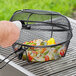A hand using an Outset non-stick grill basket to cook vegetables on a grill.