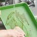 A person washing a Baker's Mark green aluminum sheet tray with a sponge.