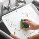 A hand using a sponge to clean a white Baker's Mark aluminum sheet pan.