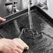 A person washing a Baker's Mark black aluminum tray in a sink with a sponge.