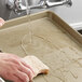 A hand holding a sponge washing a Baker's Mark non-stick aluminum sheet pan under running water.