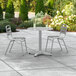 A Lancaster Table & Seating round outdoor table with metal legs and two silver chairs on a patio.