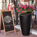 A Mayne Modesto black planter with pink flowers on an outdoor patio table.
