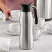 A silver and black stainless steel coffee carafe on a table with white cups.