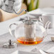 A silver kettle pouring liquid into a glass teapot with a stainless steel infuser.