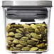An OXO Good Grips clear square plastic food storage container with green cardamom pods inside and a stainless steel lid.
