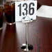 A Tablecraft stainless steel harp menu holder with a number on it sitting on a table.
