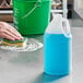 A person cleaning a school kitchen counter with a blue 1/2 gallon translucent HDPE jug of blue liquid using a sponge.