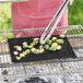 A person grilling vegetables on a Mr. Bar-B-Q grill mat over a grill using tongs.