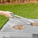 A hand holding Backyard Pro stainless steel grilling tongs to meat on a grill.