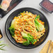 A black Acopa Izumi melamine coupe plate with noodles and vegetables and chopsticks on a table.
