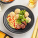 A plate of food with noodles, meat, and vegetables on an Acopa Izumi matte black melamine plate.