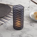 A blue frosted glass cylinder candle holder with a black spiral design on a table with a lit candle.