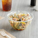 A clear plastic rose bowl filled with pasta and salad with a lid on a table.