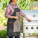A woman wearing a Backyard Pro black apron holding tongs next to a grill.