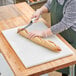 A person cutting a baguette on a white polyethylene cutting board.