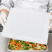 A person in white gloves holding a Vollrath Super Pan V lid over a white tray of food.