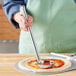 A person holding a Choice stainless steel flat bottom ladle over a pizza.