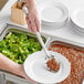 A hand using a Choice 15" Perforated Stainless Steel Basting Spoon to serve broccoli onto a white plate.