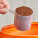 A hand using a 70 cc polypropylene scoop to fill a measuring cup with brown granules.