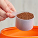 A hand using a 90 cc polypropylene scoop to pour brown granules into a container.