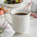 A white Acopa Condesa scalloped porcelain mug filled with coffee on a table with fruit.