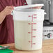 A woman using a measuring cup to pour liquid into a large white Choice food storage container with a lid.