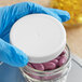 A gloved hand holding a 70/400 white ribbed plastic cap on a jar of purple pills.