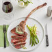 A Rastelli's bone-in tomahawk steak on a plate with asparagus and a glass of wine.