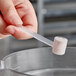 A hand holding a 2.5 cc polypropylene scoop with white powder inside a plastic tube.