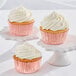 Three Enjay rose foil cupcakes with white frosting on white pedestals.
