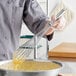 A person using a Choice stainless steel French whisk to mix yellow liquid in a bowl.